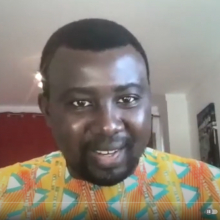A Black man in a brightly-colored shirt on a video screen talking to the camera.